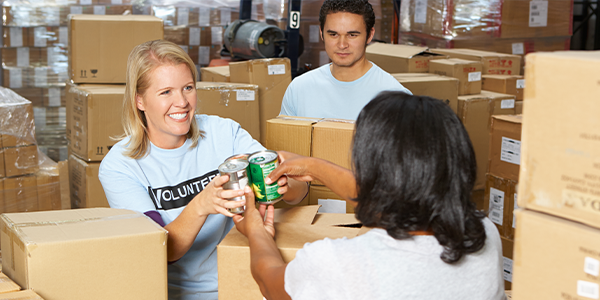Surrounded by boxes, a woman wearing a volunteer t-shirt is holding two food cans. Two other people are present wearing a similar t-shirt.
