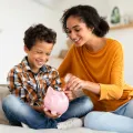 Mother and young son sitting on bed placing money in piggy bank.